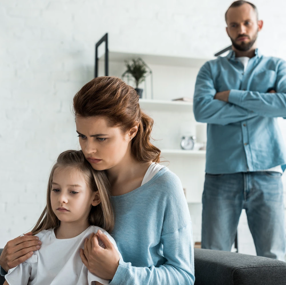 Photo of mother hugging young girl with upset father in the background